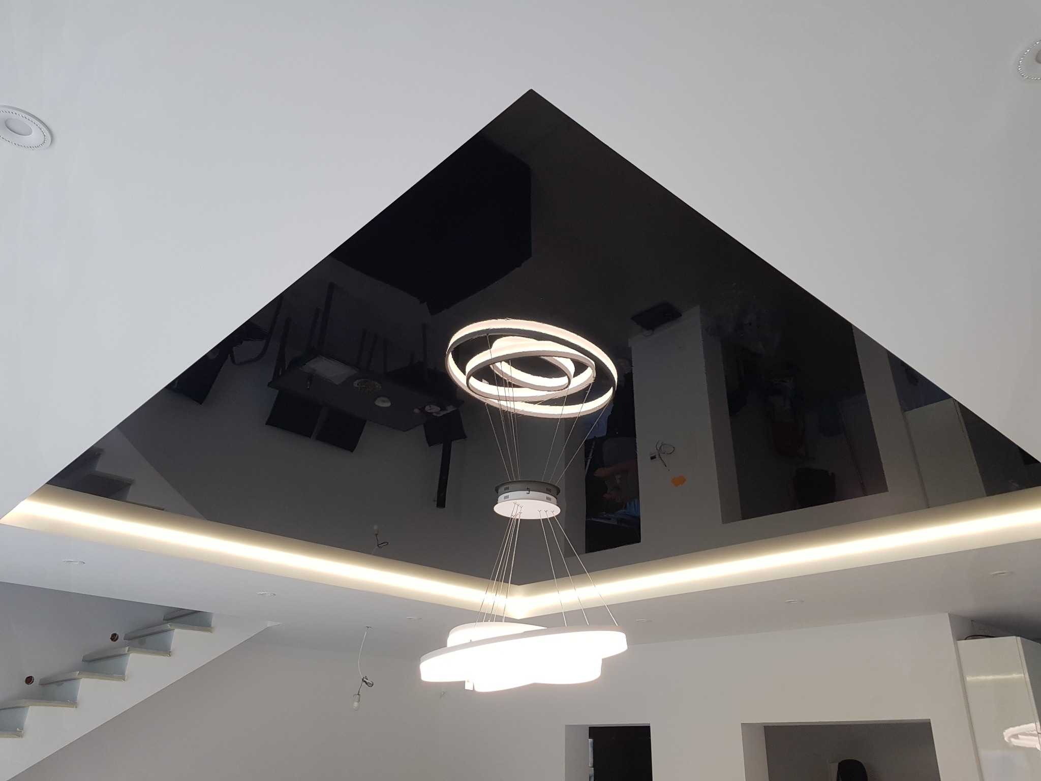Czarny, błyszczący sufit napinany w kształcie trójkąta z wbudowanym oświetleniem LED i odbijającym się w nim nowoczesnym żyrandolem, widok z dołu.