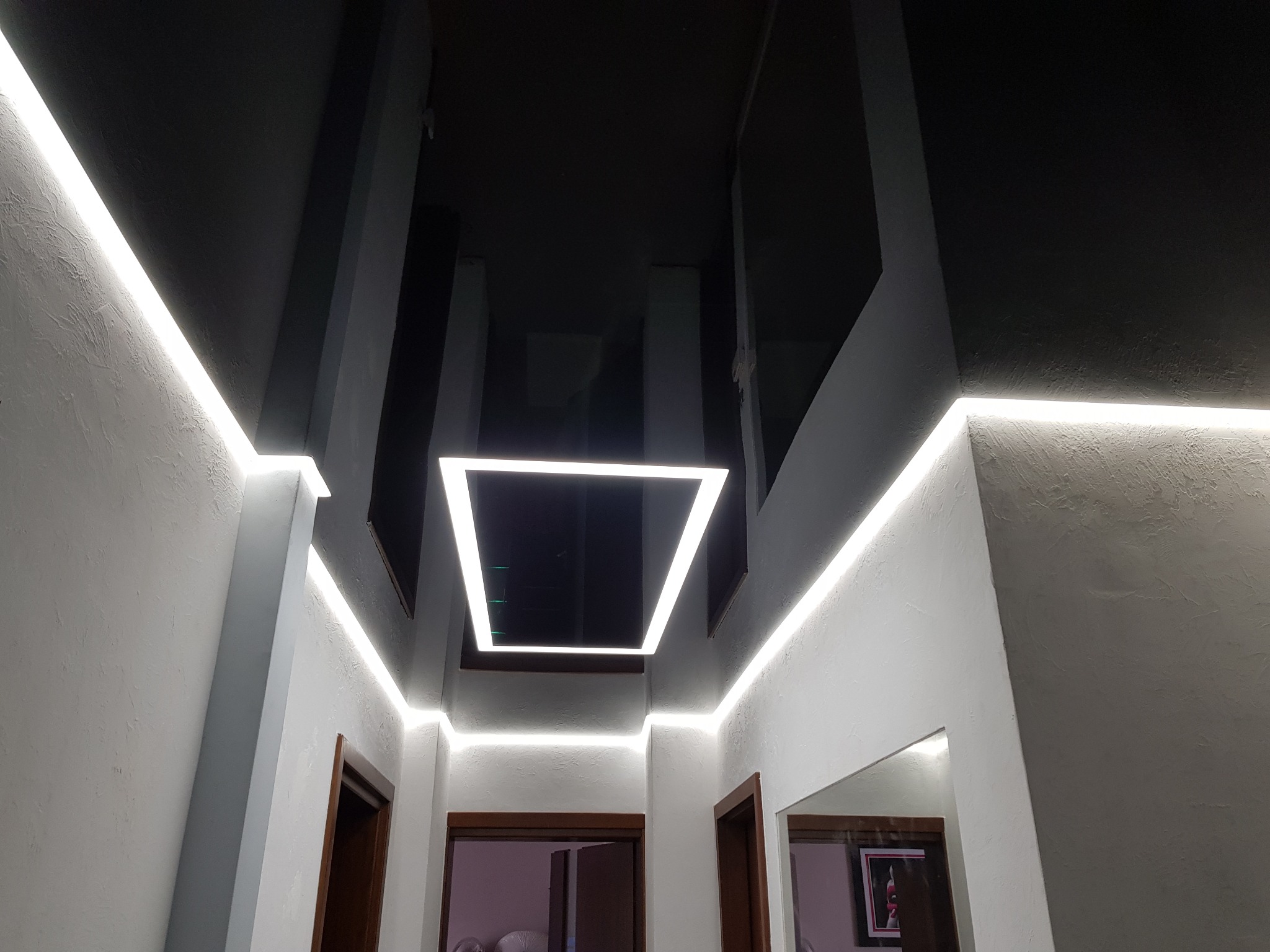 Nowoczesne wnętrze z czarnym sufitem i białymi ścianami, oświetlenie LED wzdłuż ścian i kwadratowa lampa na suficie, widoczne drzwi z ciemnego drewna.