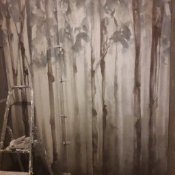 USŁUGI BUDOWLANE - Ściana z ręcznie malowanym motywem leśnym w odcieniach szarości i beżu, widoczna drabina malarska z rękawicami roboczymi i fragment podłogi w trakcie prac wykończeniowych.