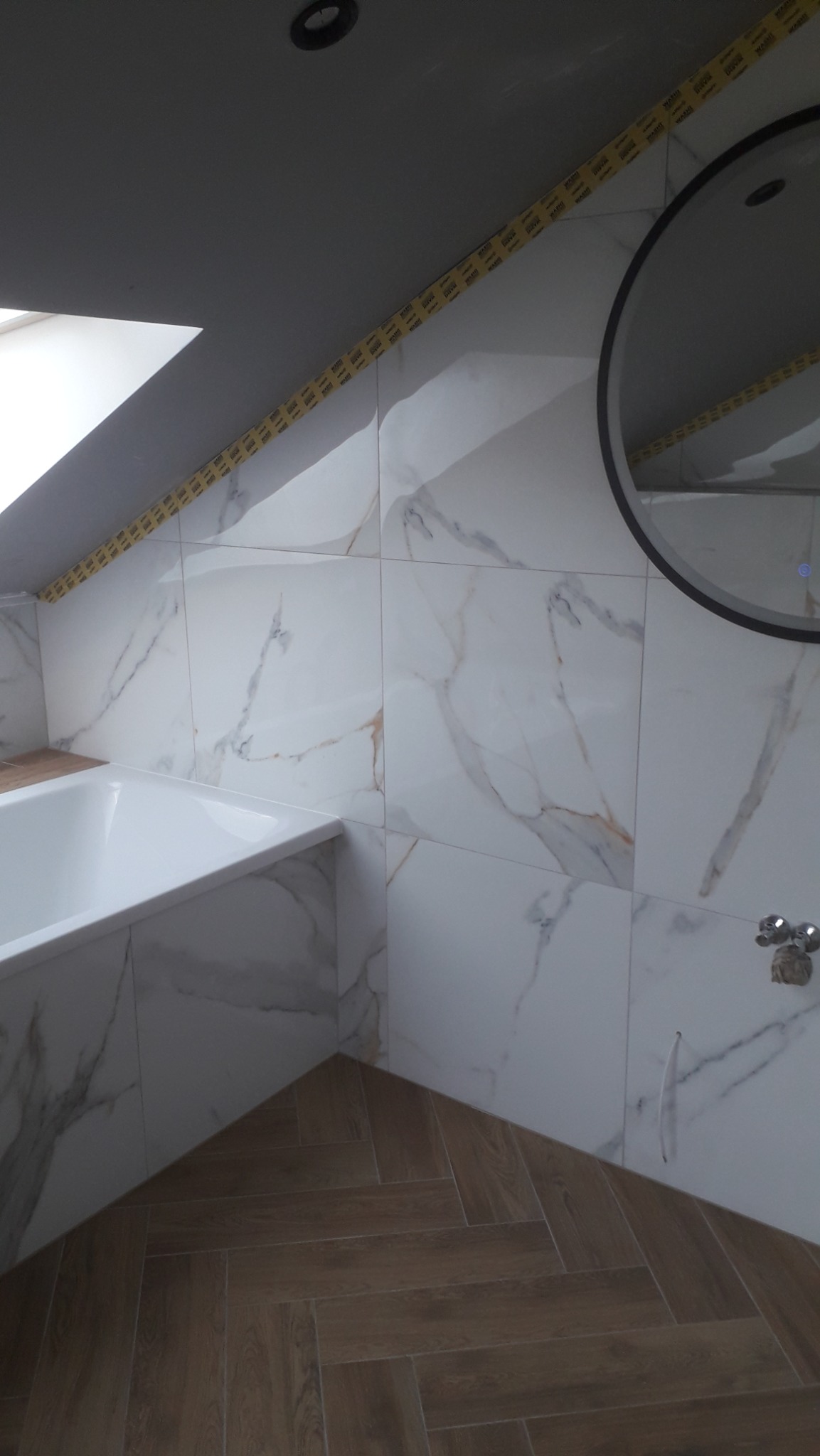Ukończona łazienka na poddaszu z białą wanną, marmurowymi płytkami na ścianach, drewnopodobną podłogą ułożoną w jodełkę i okrągłym lustrem; widoczna taśma malarska zabezpieczająca krawędzie płytek.