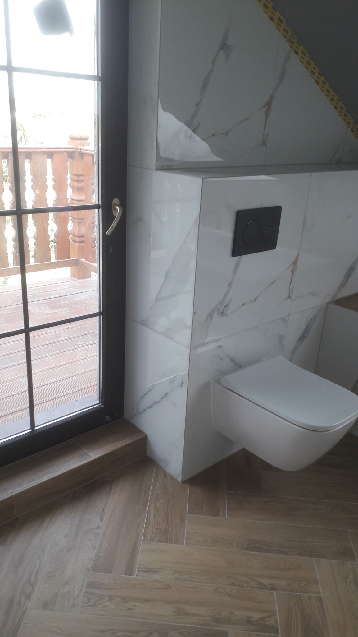 Nowoczesna toaleta podwieszana z czarnym przyciskiem spłukującym na ścianie wyłożonej białymi płytkami imitującymi marmur, podłoga z paneli ułożonych w jodełkę, widok na drewniany taras przez okno.