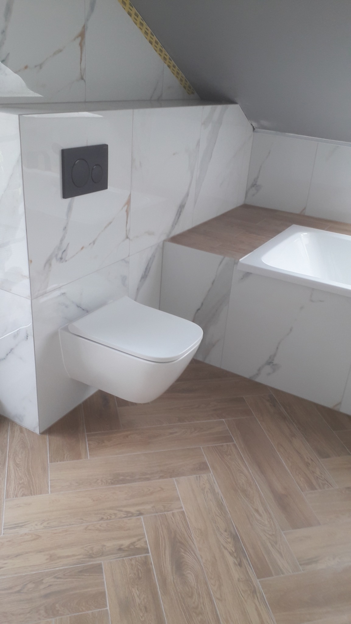 Nowoczesna łazienka na poddaszu z podwieszaną toaletą, białymi płytkami imitującymi marmur na ścianach i drewnopodobną podłogą ułożoną w jodełkę.