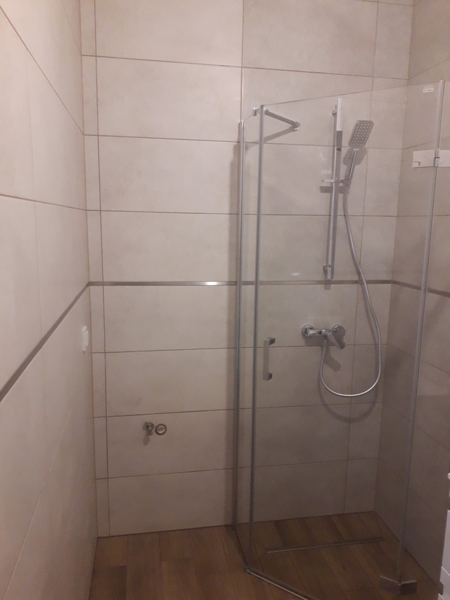 Kabina prysznicowa z chromowaną armaturą i deszczownicą, wykończona płytkami w odcieniach beżu z poziomym, metalowym akcentem, na podłodze drewnopodobne płytki.