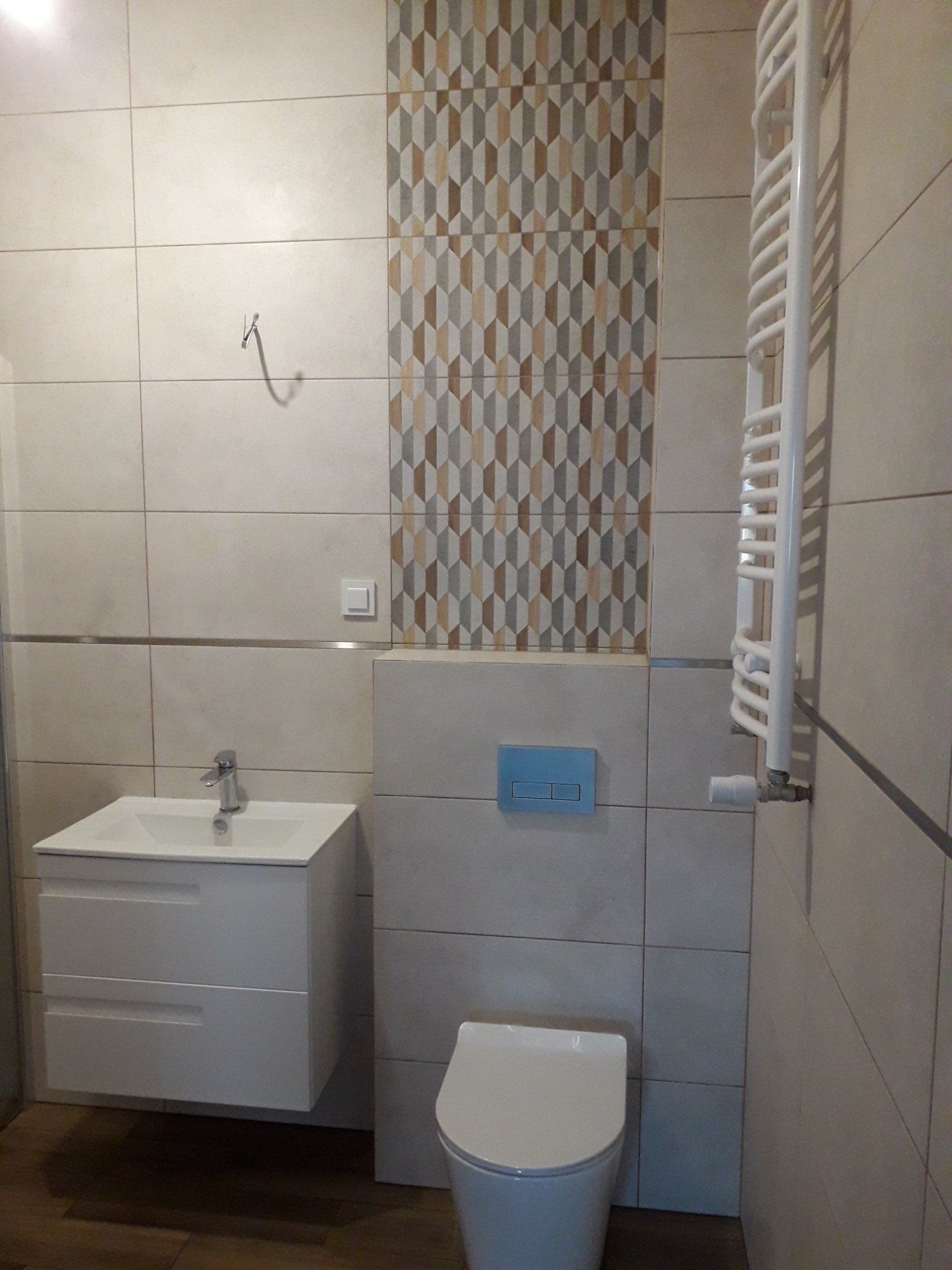 Wykończona łazienka z beżowymi płytkami, dekoracyjnym pasem płytek w geometryczny wzór, białą wiszącą szafką z umywalką, toaletą i grzejnikiem drabinkowym