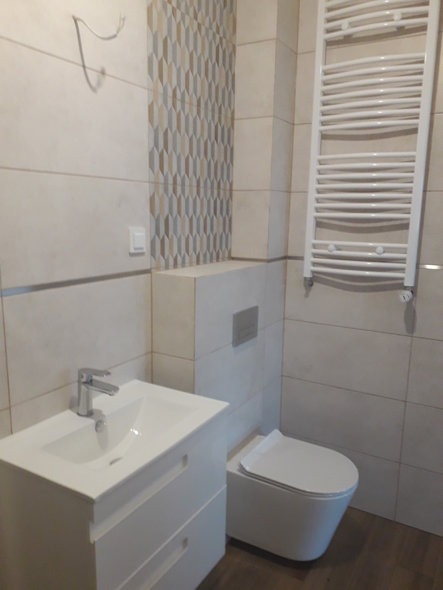 Wykończona łazienka we Wrocławiu z białymi płytkami, umywalką z szafką, podwieszaną toaletą i grzejnikiem drabinkowym. Uwagę przyciąga dekoracyjna mozaika na ścianie.