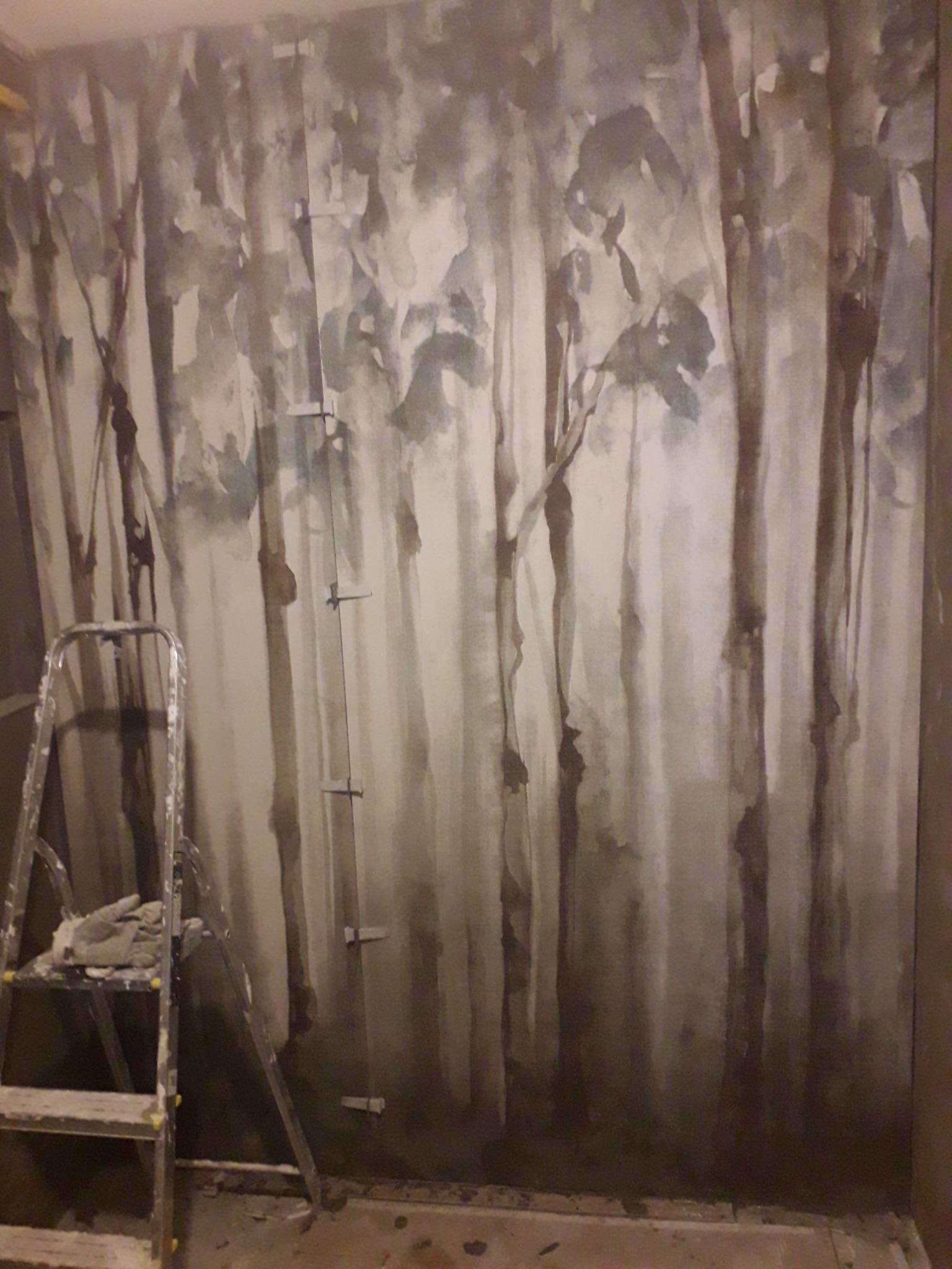 Ściana z ręcznie malowanym motywem leśnym w odcieniach szarości i beżu, widoczna drabina malarska z rękawicami roboczymi i fragment podłogi w trakcie prac wykończeniowych.