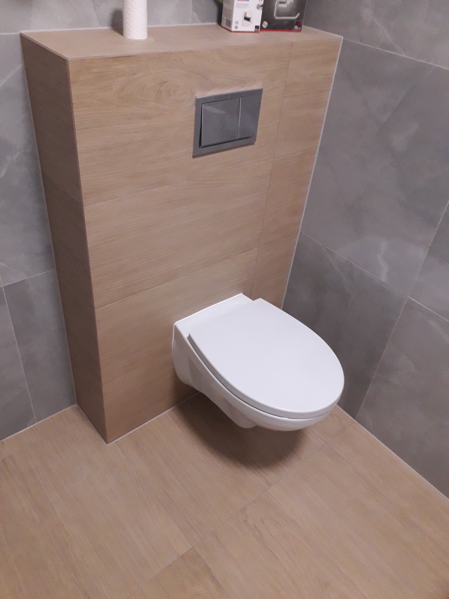 Nowoczesna, podwieszana toaleta z przyciskiem spłukującym w obudowie z płytek imitujących drewno, na tle szarych płytek ściennych i podłogowych.