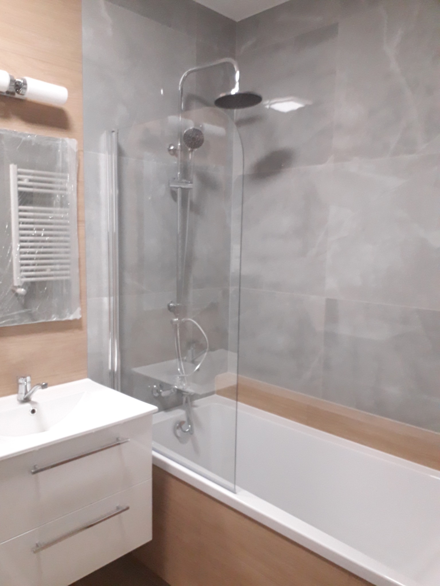 Nowoczesna łazienka z wanną, szklanym parawanem nawannowym, białą umywalką z szafką i grzejnikiem drabinkowym na ścianie z drewnopodobnymi panelami.