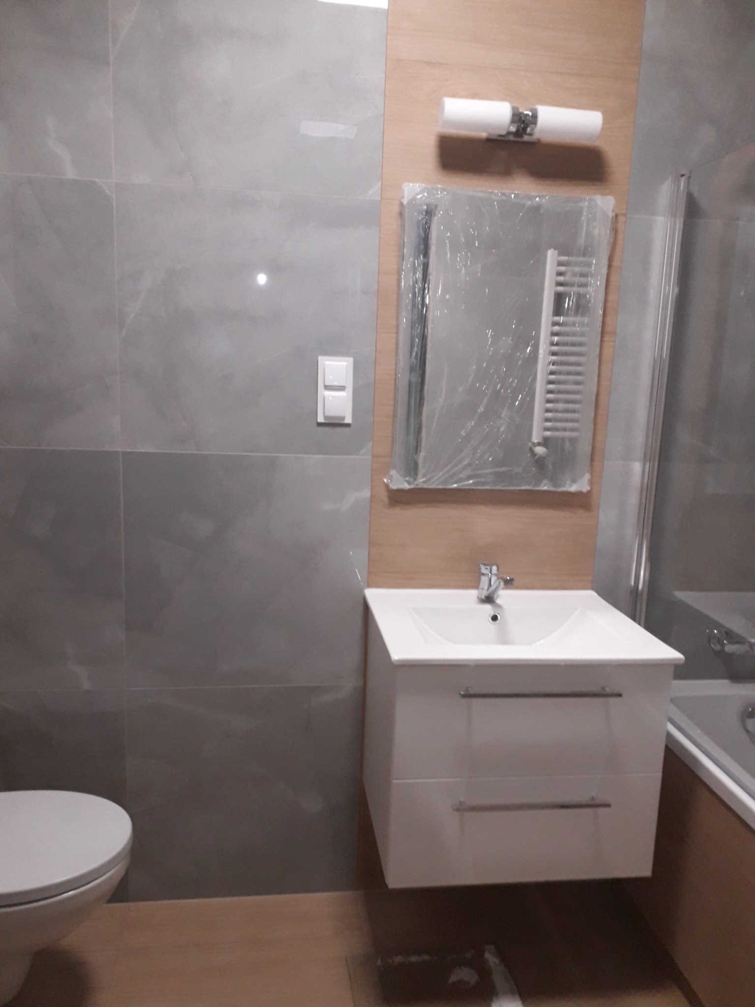 Nowoczesna łazienka w szarościach i drewnie: umywalka z szafką, lustro w folii ochronnej z grzejnikiem drabinkowym, płytki imitujące marmur i drewnopodobna okładzina. W tle widoczny fragment toalety.