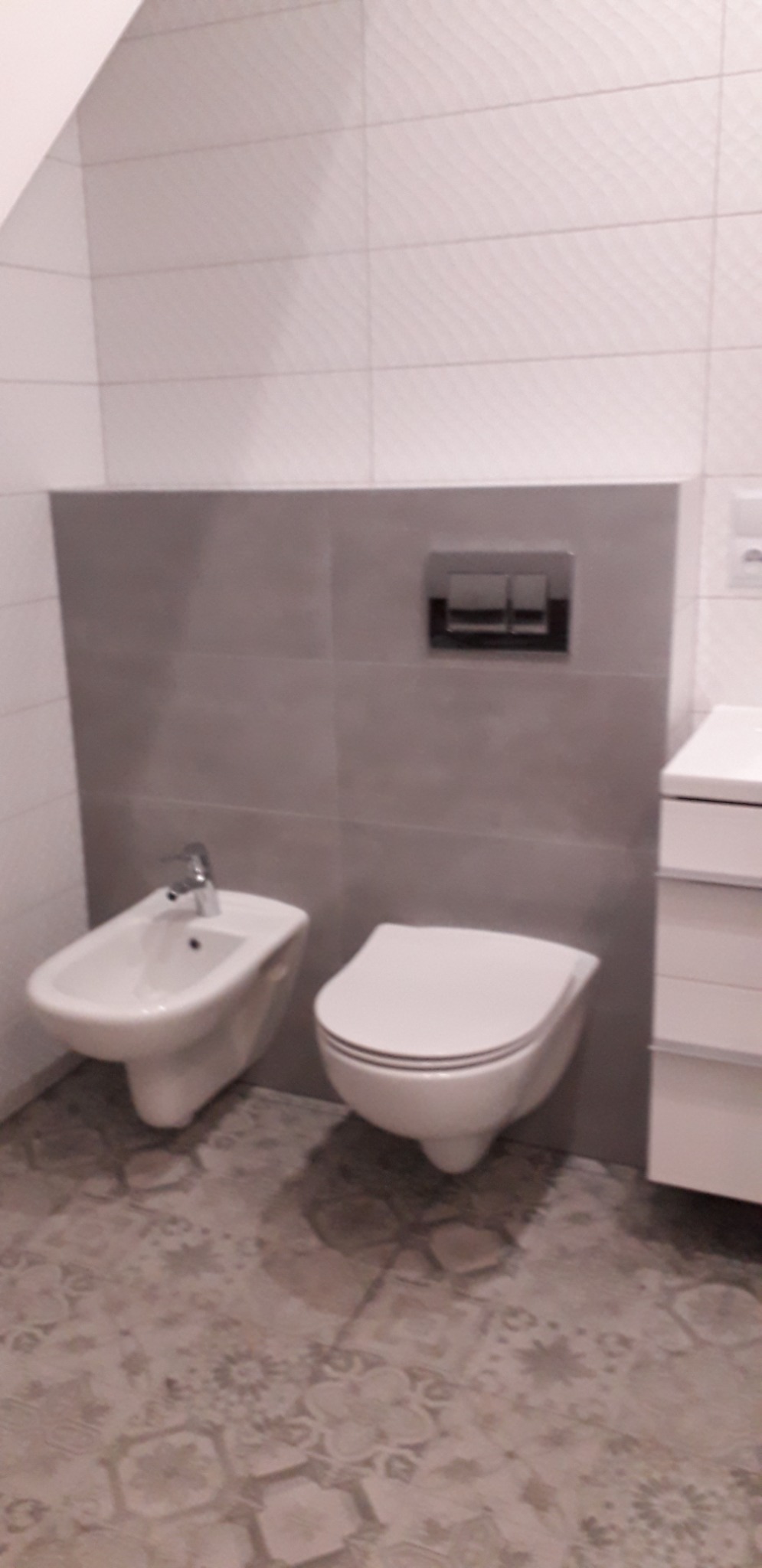 Nowoczesna łazienka z wiszącą toaletą i bidetem na tle szarych płytek, z wzorzystą podłogą i chromowanym przyciskiem spłuczki.