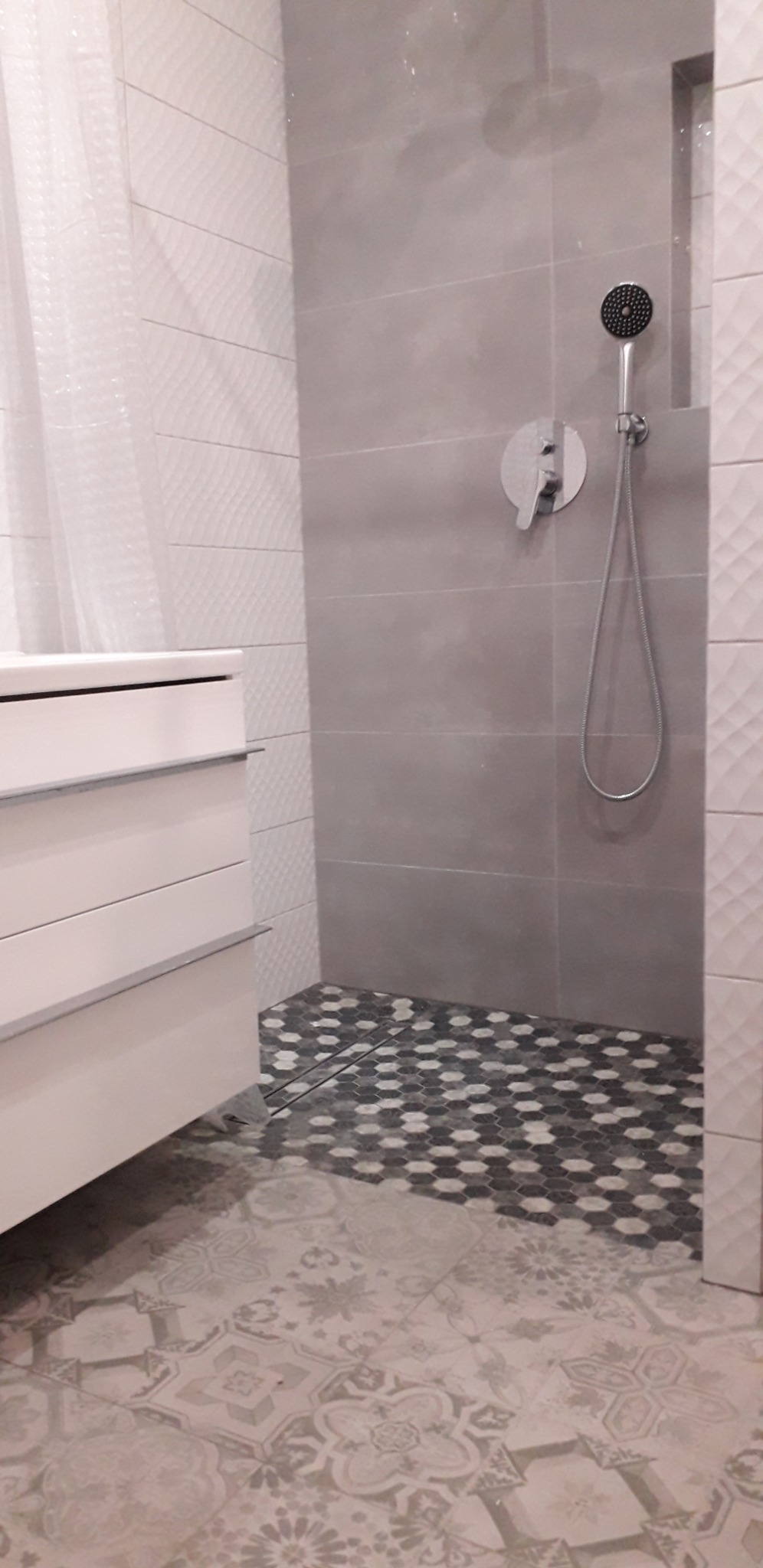 Nowoczesna łazienka z białą szafką umywalkową, szarą kabiną prysznicową z deszczownicą i mozaikową podłogą w strefie prysznica, przechodzącą w patchworkowe płytki podłogowe.