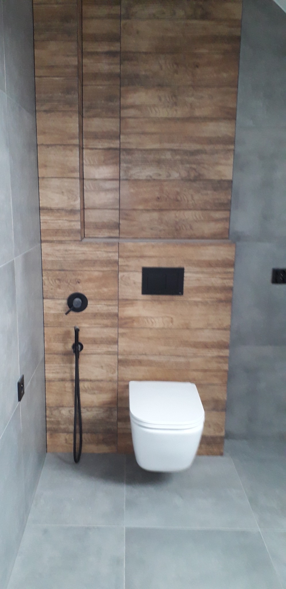 Nowoczesna łazienka z podwieszaną toaletą, drewnopodobnymi płytkami na ścianie i szarą podłogą. Na ścianie widoczny czarny panel sterujący spłuczką oraz czarny bidetta.