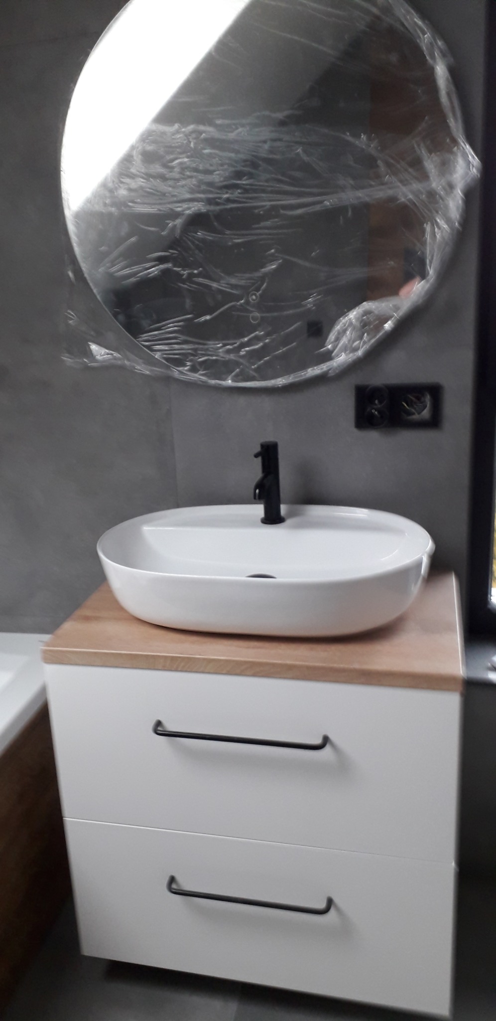 Nowoczesna łazienka z białą szafką pod umywalkę z drewnianym blatem i okrągłym lustrem w folii ochronnej, czarna bateria umywalkowa i szara ściana z gniazdkiem.