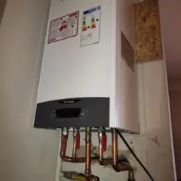 Nowoczesny piec gazowy Ariston Clas X, zamontowany na ścianie, z widocznymi miedzianymi rurami i zaworami, na tle ściany z widoczną fakturą i fragmentem płyty OSB.