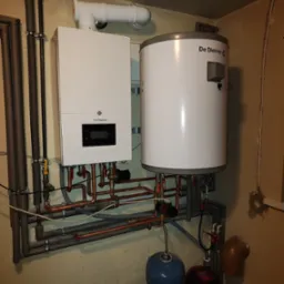 Biały, wiszący piec gazowy De Dietrich z podłączonym zasobnikiem ciepłej wody, widoczne rury miedziane i instalacja, ściana w odcieniach beżu.