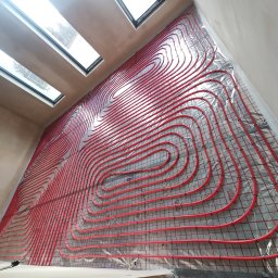 JJ Systemy Grzewcze Sp. z o.o. - Czerwone rury ogrzewania podłogowego ułożone spiralnie na srebrnej macie izolacyjnej w jasnym pomieszczeniu z oknami dachowymi. Widok z góry pod kątem.