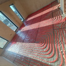 JJ Systemy Grzewcze Sp. z o.o. - Czerwone rury ogrzewania podłogowego ułożone na folii, widok z góry, w tle okna w surowym wnętrzu. Instalacja przed wylaniem posadzki.