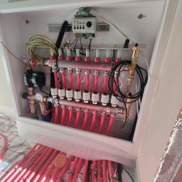 JJ Systemy Grzewcze Sp. z o.o. - Rozdzielacz ogrzewania podłogowego w białej szafce, widoczne czerwone rury, manometry i zawory. Instalacja w trakcie montażu, widoczne elementy mocujące rury.