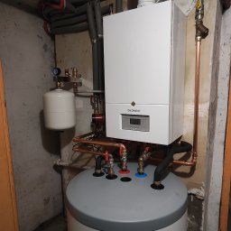 JJ Systemy Grzewcze Sp. z o.o. - Biały, wiszący piec gazowy De Dietrich z szarym zbiornikiem na wodę w kotłowni. Widoczne miedziane rury i zawory. Ściany z surowego betonu.