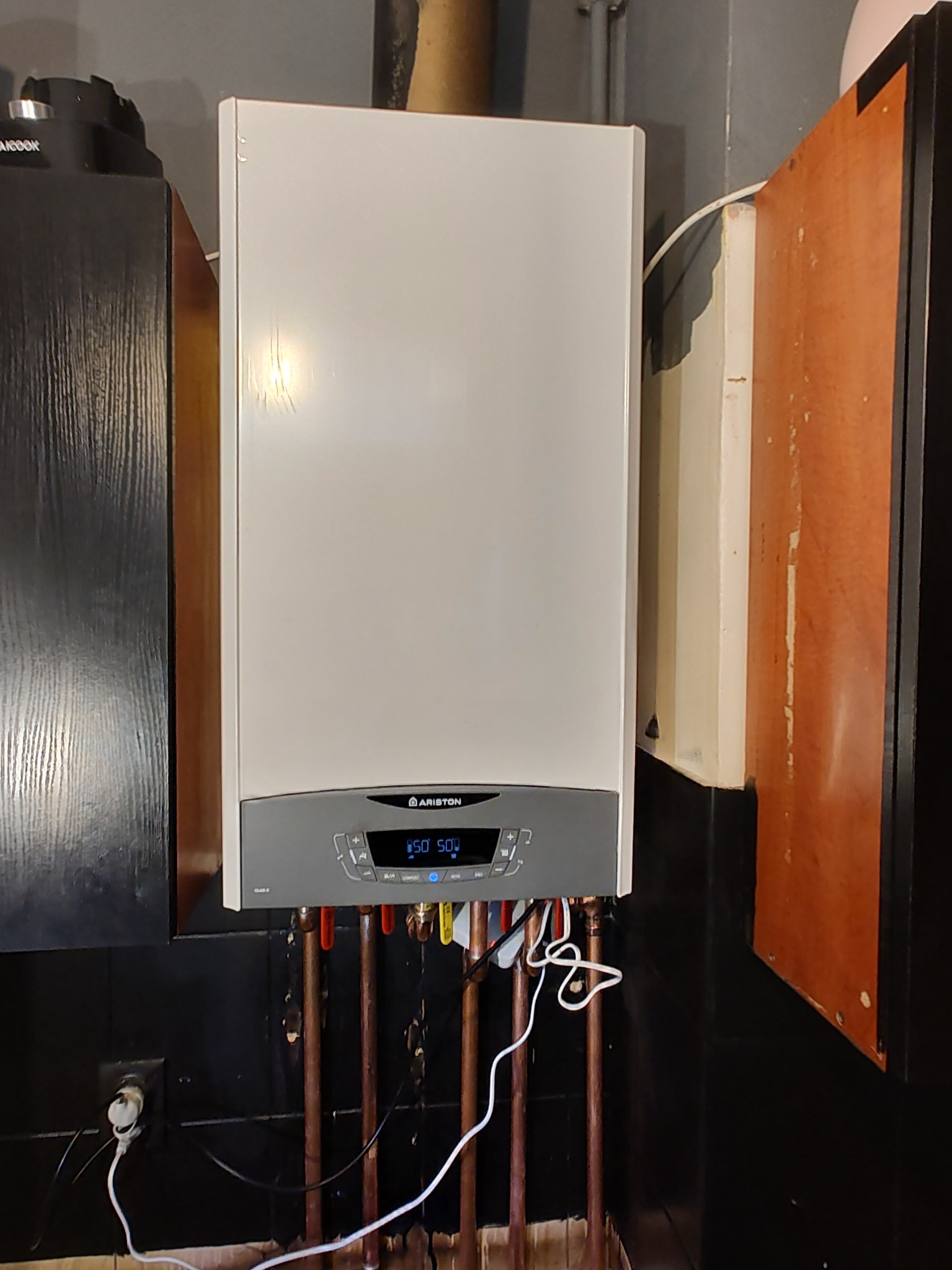 Biały, wiszący piec gazowy marki Ariston z widocznymi rurami miedzianymi i przewodami elektrycznymi, zamontowany na ciemnej ścianie z widocznym gniazdkiem elektrycznym.