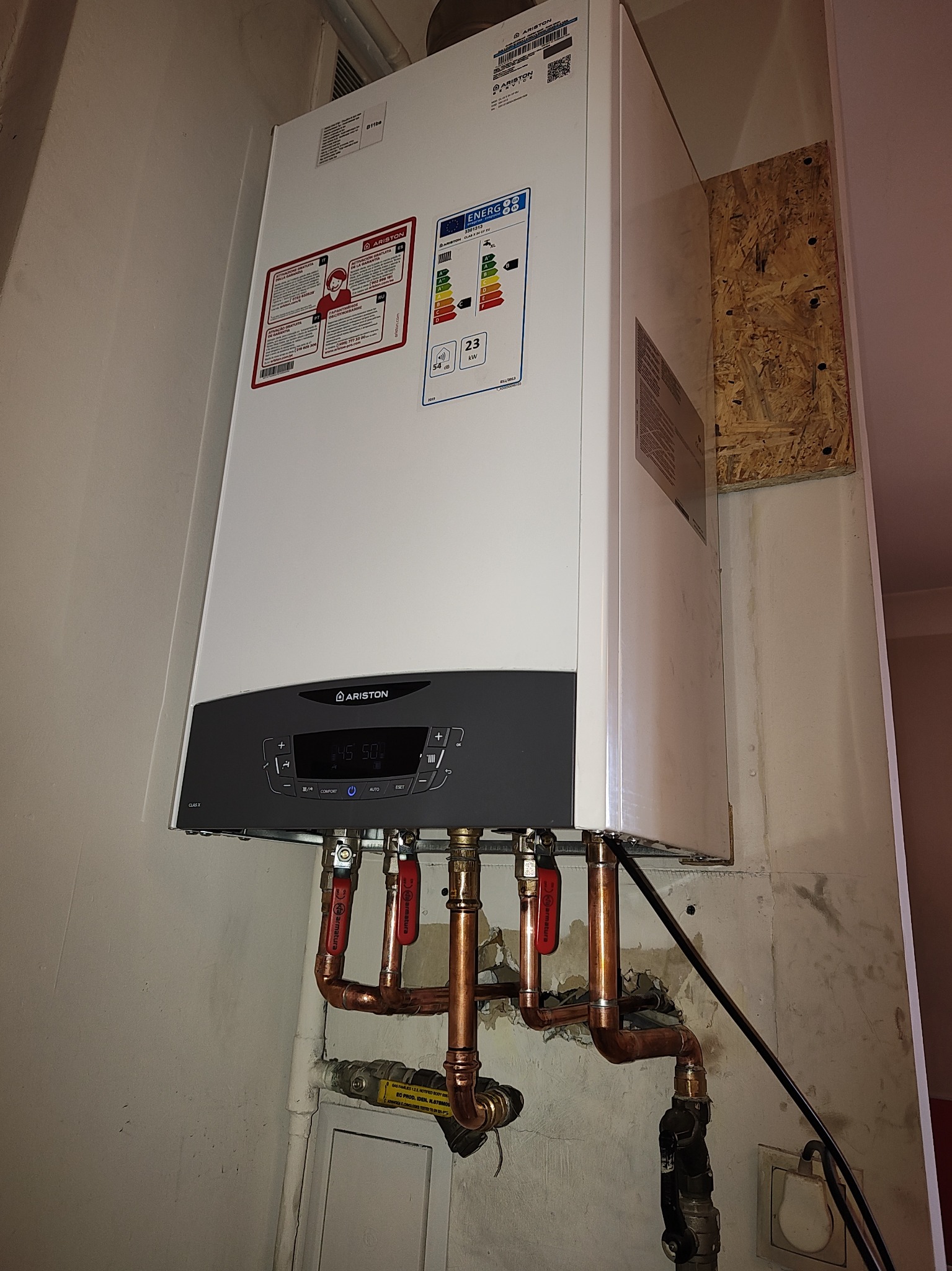 Nowoczesny piec gazowy Ariston Clas X, zamontowany na ścianie, z widocznymi miedzianymi rurami i zaworami, na tle ściany z widoczną fakturą i fragmentem płyty OSB.