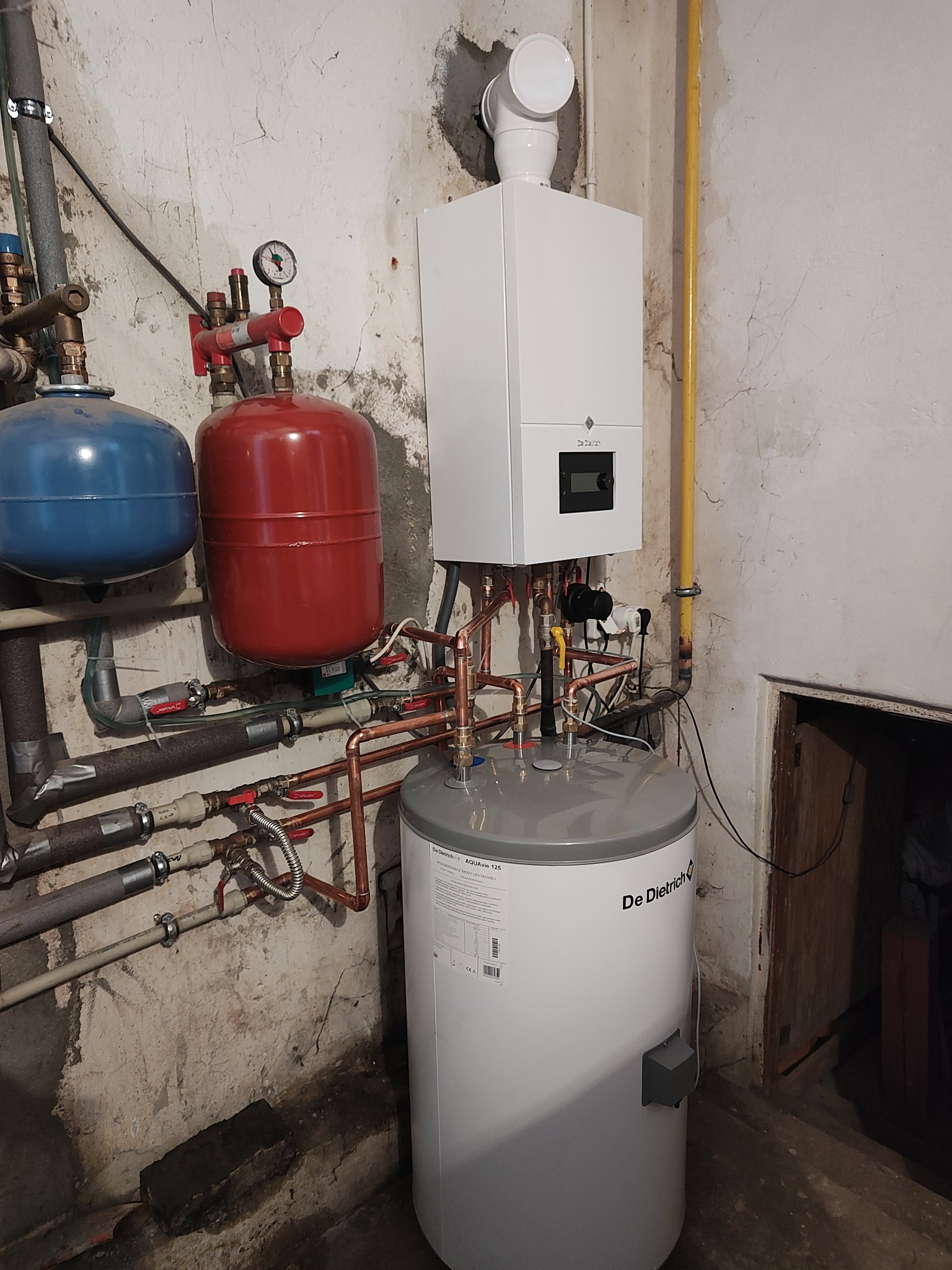 Nowoczesny piec gazowy De Dietrich z zasobnikiem ciepłej wody i instalacją hydrauliczną w piwnicy o surowych ścianach.