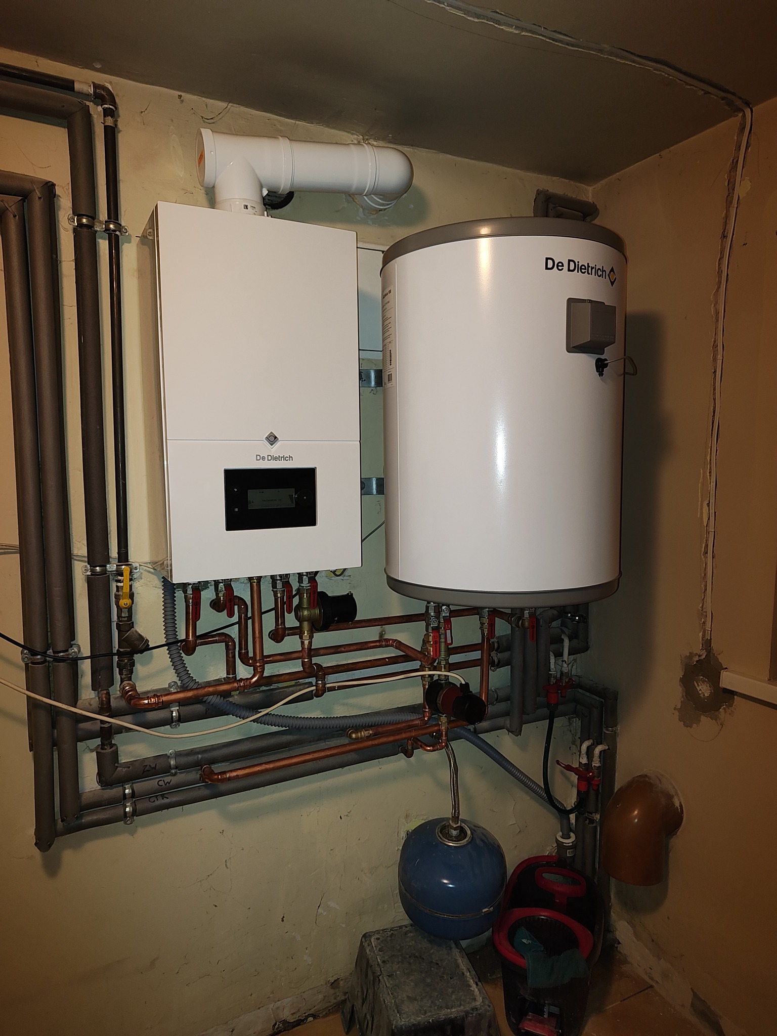 Biały, wiszący piec gazowy De Dietrich z podłączonym zasobnikiem ciepłej wody, widoczne rury miedziane i instalacja, ściana w odcieniach beżu.