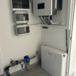 Instalacja wewnętrzna z inwerterem SOFAR, magazynem energii SOFAR, rozdzielnicami elektrycznymi i filtrem wody z licznikiem, zamontowana w pomieszczeniu o białych ścianach i szarych płytkach...