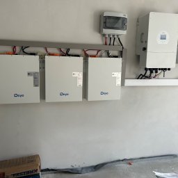 Energy 4 you Krzysztof Słomka - Instalacja magazynów energii Deye na szarej ścianie. Widoczne trzy białe moduły, inwerter i skrzynki przyłączeniowe. Surowy stan wykończenia wnętrza.