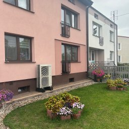 Energy 4 you Krzysztof Słomka - Klimatyzator Panasonic zamontowany na zewnątrz różowego domu, widoczny z poziomu trawnika z kwiatami. Ujęcie z boku, z widocznym fragmentem sąsiedniego budynku.