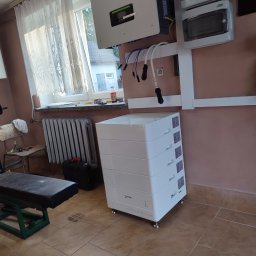 Energy 4 you Krzysztof Słomka - Biały magazyn energii marki Nuras na brązowej podłodze, obok grzejnika i okna z firanką. Na ścianie falownik i skrzynki elektryczne. Domowa siłownia w tle.