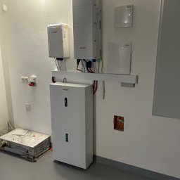 Energy 4 you Krzysztof Słomka - Biały magazyn energii z podłączonymi przewodami, zamontowany na ścianie w pomieszczeniu. Widoczne skrzynki elektryczne i gniazda. Na podłodze leżą płyty.