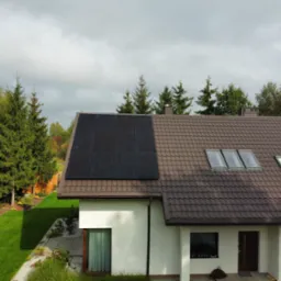 Dom jednorodzinny z brązowym dachem i panelami fotowoltaicznymi na połowie dachu. Widok z góry, w tle drzewa i pochmurne niebo.