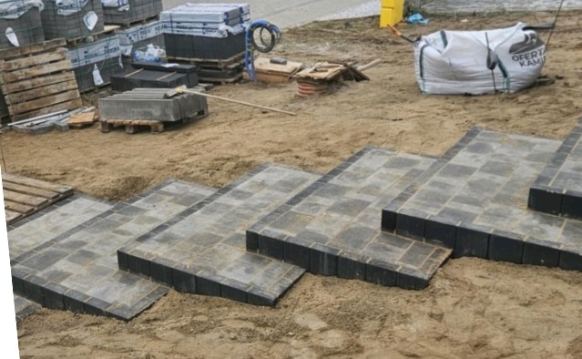 Schody z kostki brukowej w Gryfinie, częściowo ułożone na piasku. W tle palety z materiałami budowlanymi i worki. Widok z dołu, ukazujący strukturę i kolorystykę.