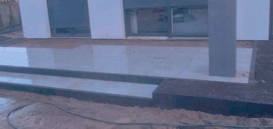 Wyłożony jasnymi płytkami taras z dwustopniowymi schodami, kontrastującymi ciemnymi obrzeżami i kolumną na tle białej elewacji domu z dużymi oknami.