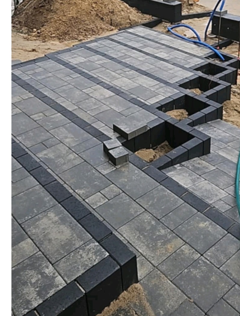 Układanie kostki brukowej w szaro-czarnej kolorystyce. Widoczne niezakończone fragmenty i piasek. Ujęcie z góry pokazuje wzór ułożenia kostki.