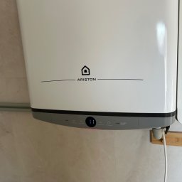 Hydro-mił Usługi Hydrauliczne - Biały, wiszący bojler Ariston z wyświetlaczem LED wskazującym temperaturę, zamontowany na marmurowej ścianie. Widoczny kabel zasilający i fragment instalacji.