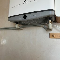 Hydro-mił Usługi Hydrauliczne - Biały, podwieszany bojler Ariston z szarym panelem sterowania, zamontowany na beżowej ścianie z widocznymi rurami doprowadzającymi wodę i elementami instalacji.