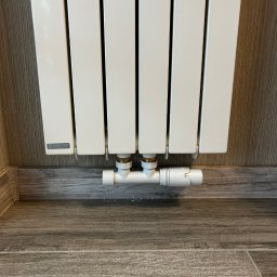 Biały grzejnik panelowy Cordivari z termostatem na tle drewnopodobnej ściany i podłogi. Widok z dołu, prezentujący estetyczne połączenie z instalacją.