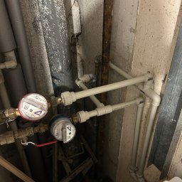 Instalacja wodna z dwoma licznikami Kalon, rurami z tworzywa sztucznego i zaworem z czerwoną rączką w surowym pomieszczeniu z popękaną ścianą.