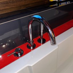 Bateria wannowa chromowana z wylewką kaskadową i panelem sterującym na tle czarno-czerwonych płytek i okna. Widoczny silikon sanitarny.