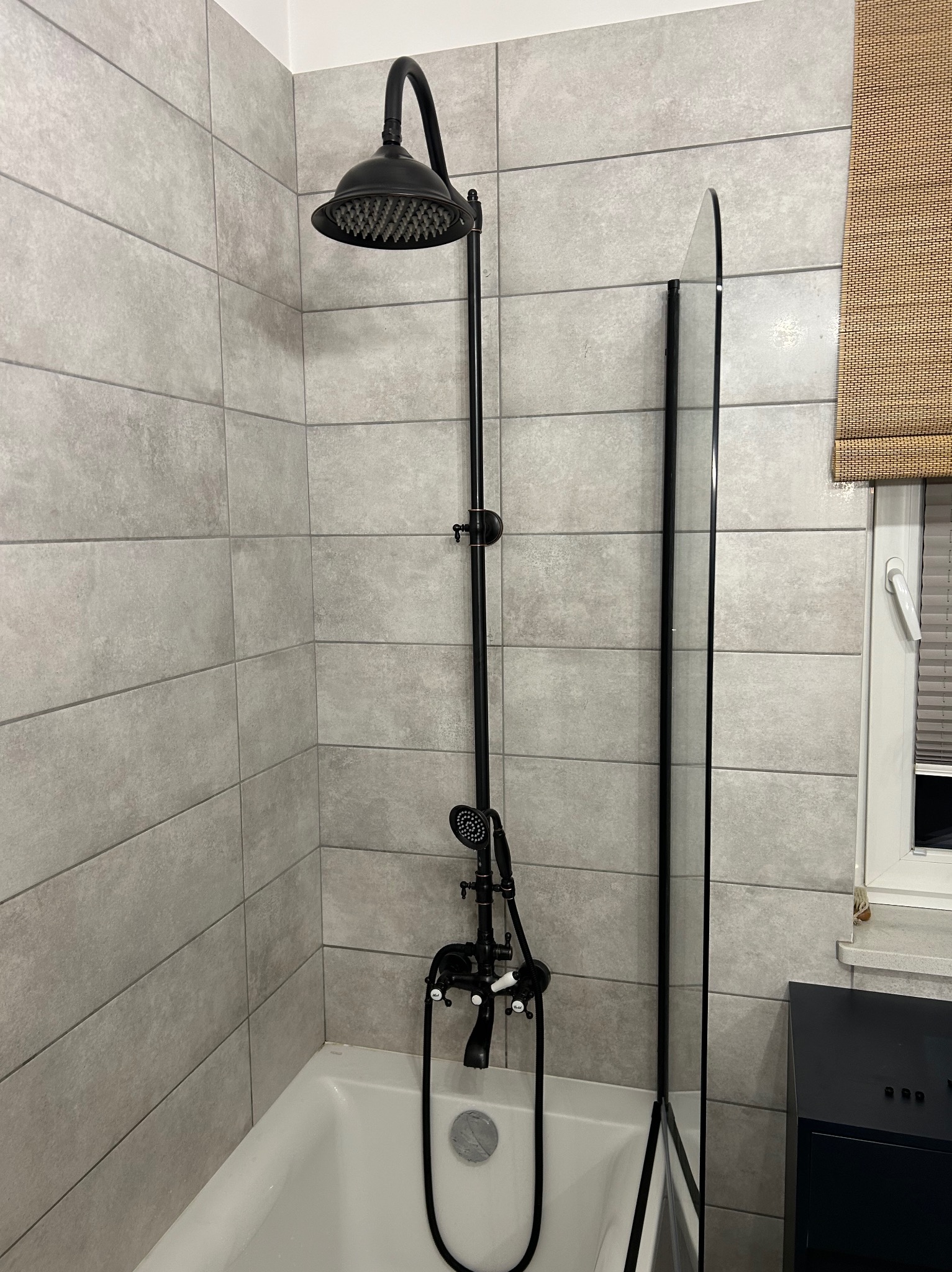 Czarna bateria prysznicowa z deszczownicą i słuchawką, zamontowana nad wanną z parawanem. Szare płytki na ścianie, roleta w oknie. Nowoczesna łazienka.