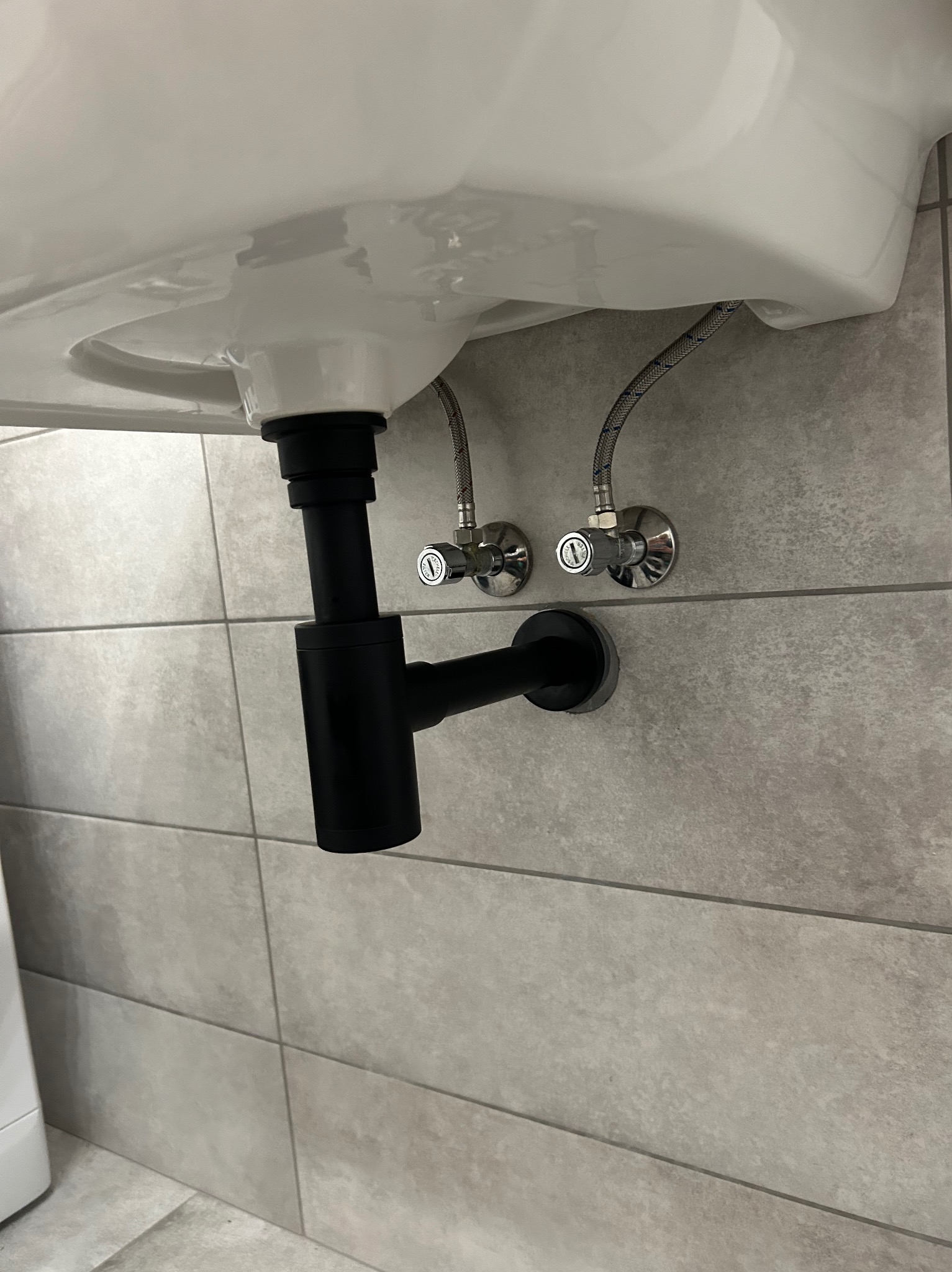 Czarny syfon umywalkowy zamontowany na szarej, kafelkowanej ścianie. Widoczne zawory dopływu wody z elastycznymi wężykami.