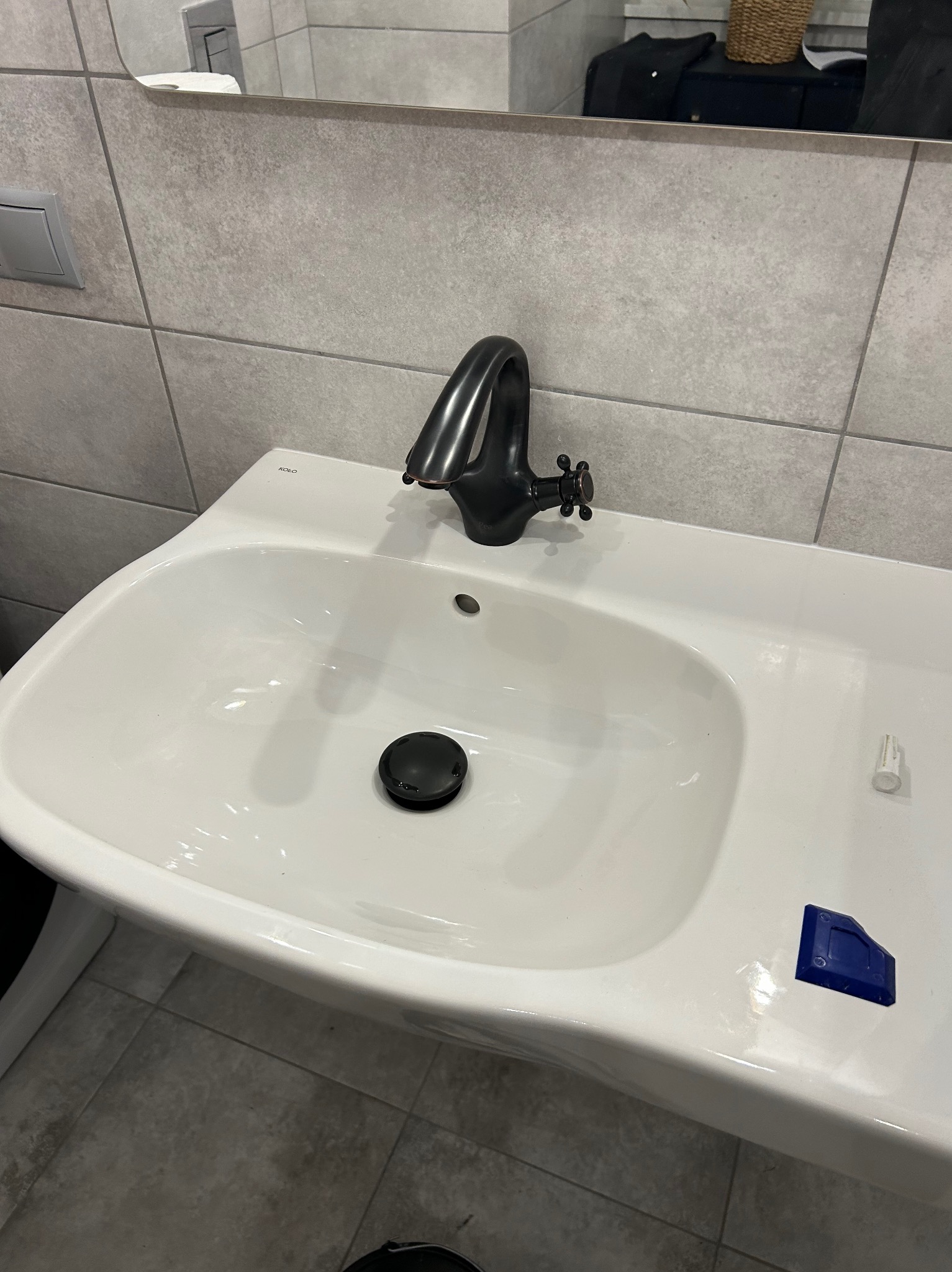 Biała umywalka KOLO z czarną armaturą, widoczne narzędzia do silikonowania. Ujęcie z góry, szare płytki w tle. Nowoczesna łazienka po montażu.