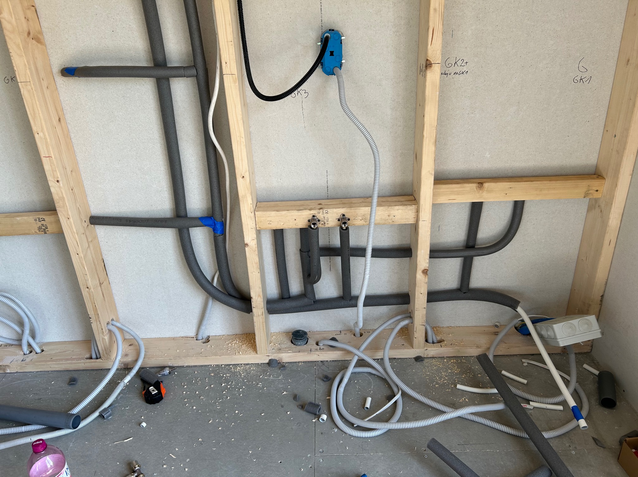 Instalacja wodna i elektryczna w ścianie szkieletowej. Rury w izolacji, przewody w peszlach, niebieska puszka elektryczna. Widoczne elementy montażowe i fragment podłogi.