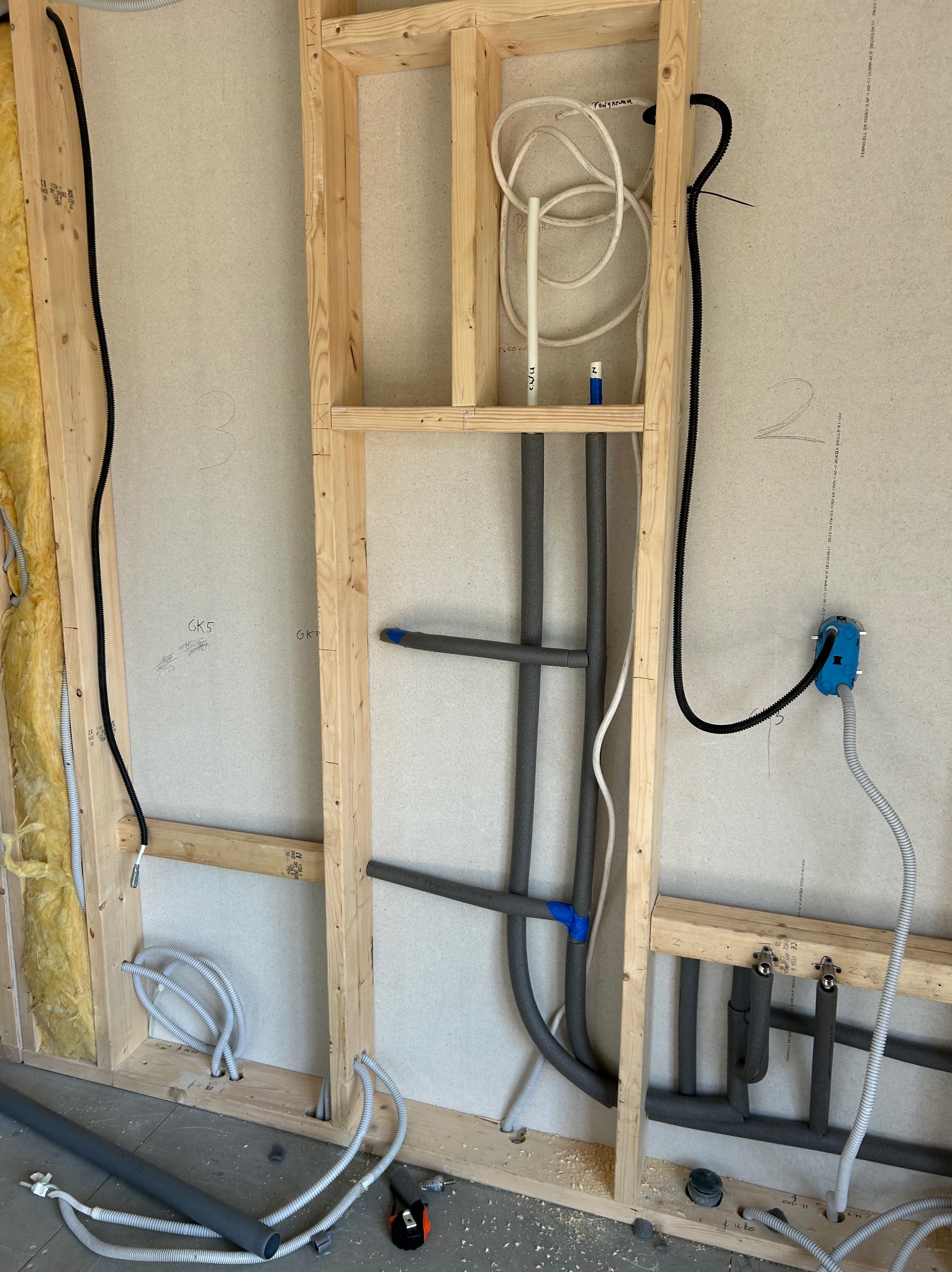Szare rury instalacji wodnej w drewnianej konstrukcji ściany działowej, widoczne przewody elektryczne i fragment izolacji. Etap prac instalacyjnych w nowym budynku.