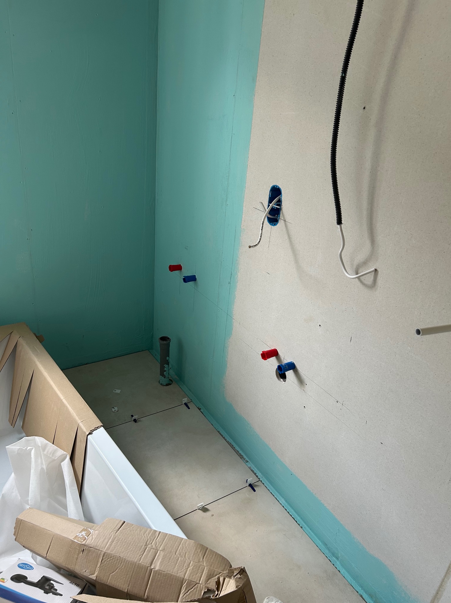 Przygotowanie łazienki do montażu wanny: surowe ściany z wyprowadzonymi przyłączami wodnymi i elektrycznymi, widoczne płytki podłogowe i zabezpieczona hydroizolacją dolna część ścian.