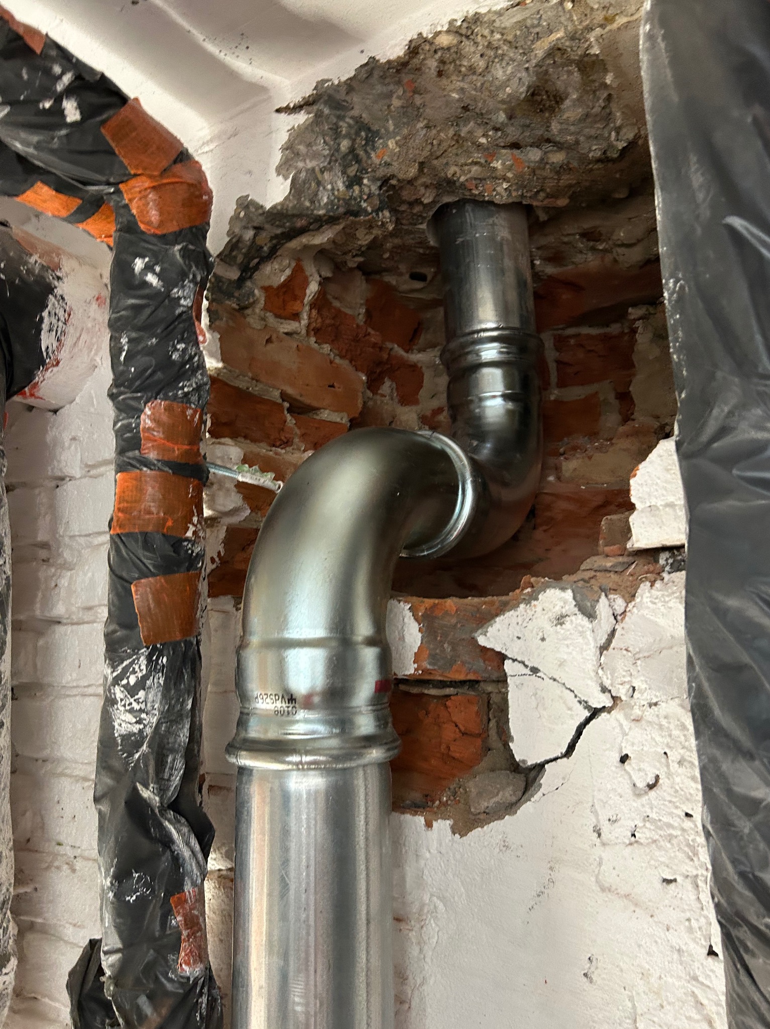 Błyszczące, metalowe rury instalacji wodnej, z widocznymi złączkami, prowadzone przez otwór w murze z cegły, częściowo otynkowanym na biało. Izolacja rur w tle.