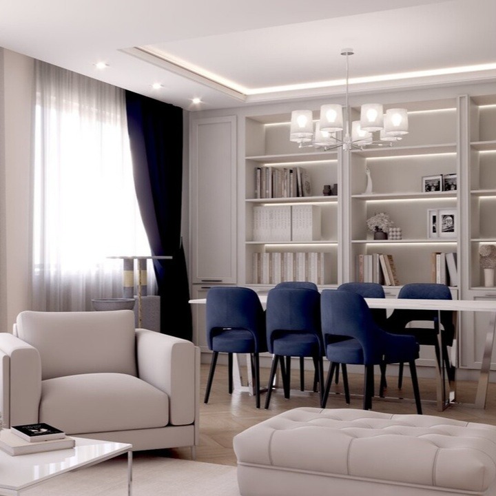 Jasne wnętrze salonu z beżową sofą, pufą i białym stolikiem kawowym, uzupełnione granatowymi krzesłami przy białym stole jadalnym, na tle regału z książkami i dekoracjami.