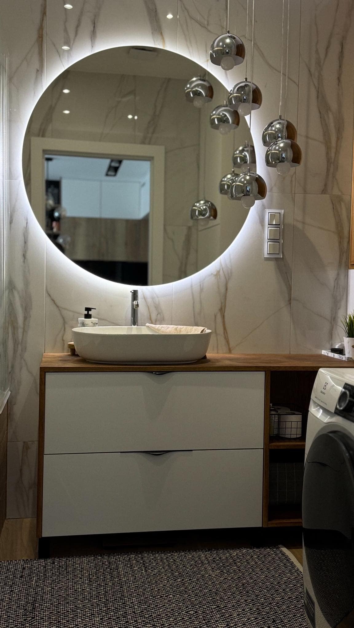 Nowoczesna łazienka z okrągłym lustrem LED, umywalką nablatową, szafką z szufladami i marmurowymi płytkami na ścianie, uzupełniona o zwisające lampy w kształcie kul i pralkę Electrolux.